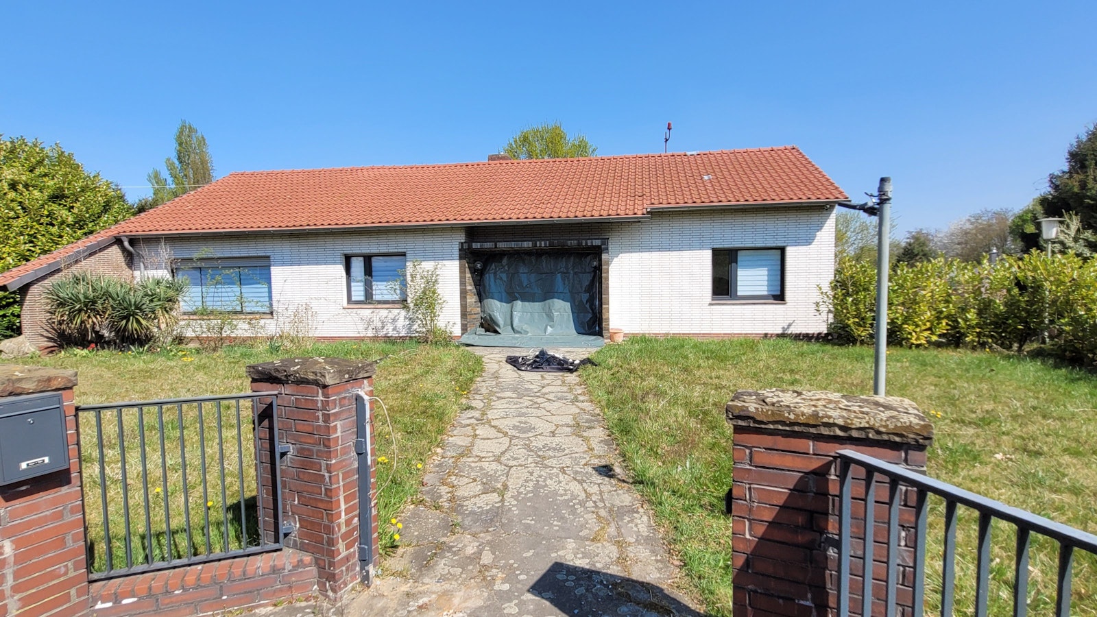 Ein in die Jahre gekommenes Haus mit heller Fassade, die Rolläden an den drei Fenstern sind heruntergelassen, dunkle Folie ersetzt die Eingangstür.