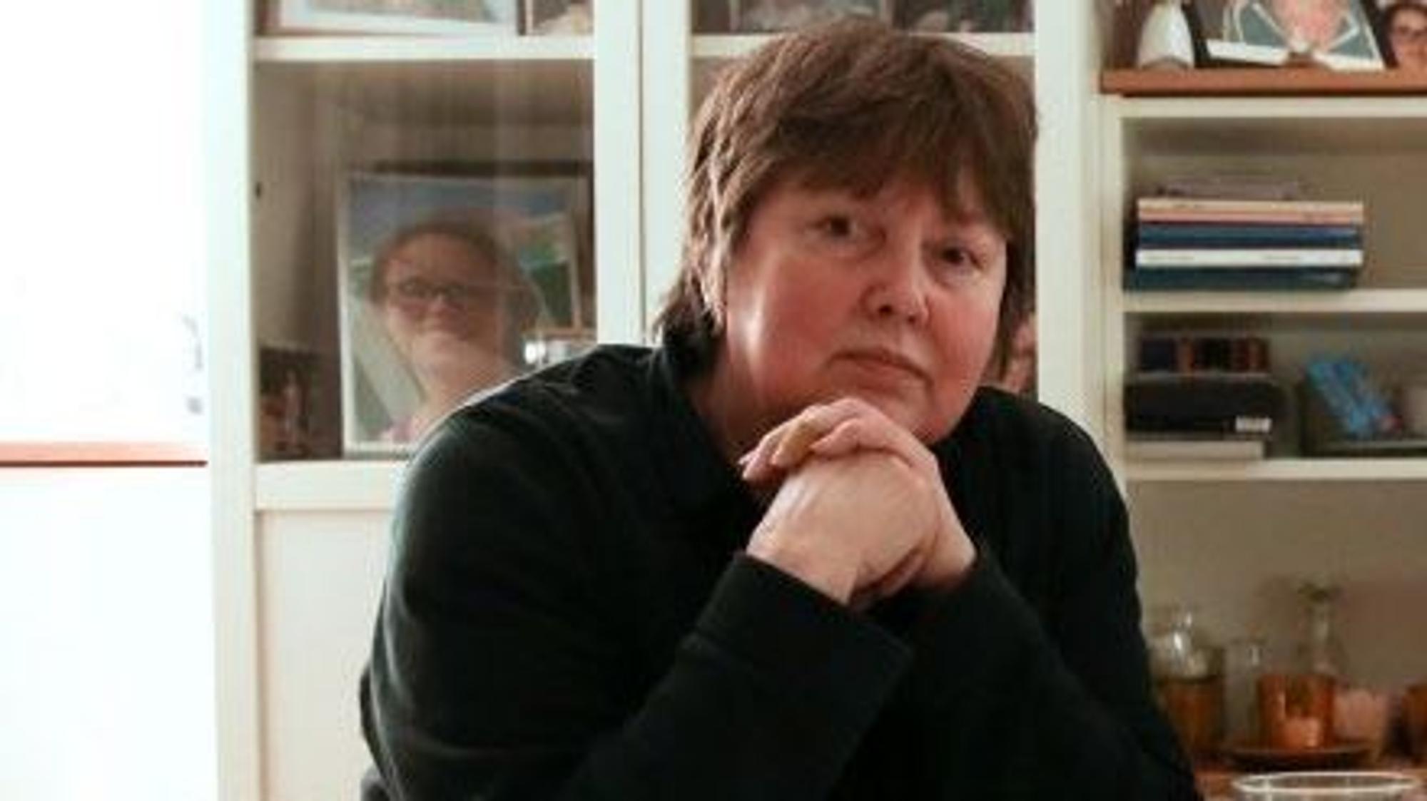Marita Scheidel stützt ihren Kopf im Sitzen leicht auf ihre Hände, die sie übereinander gelegt hat.