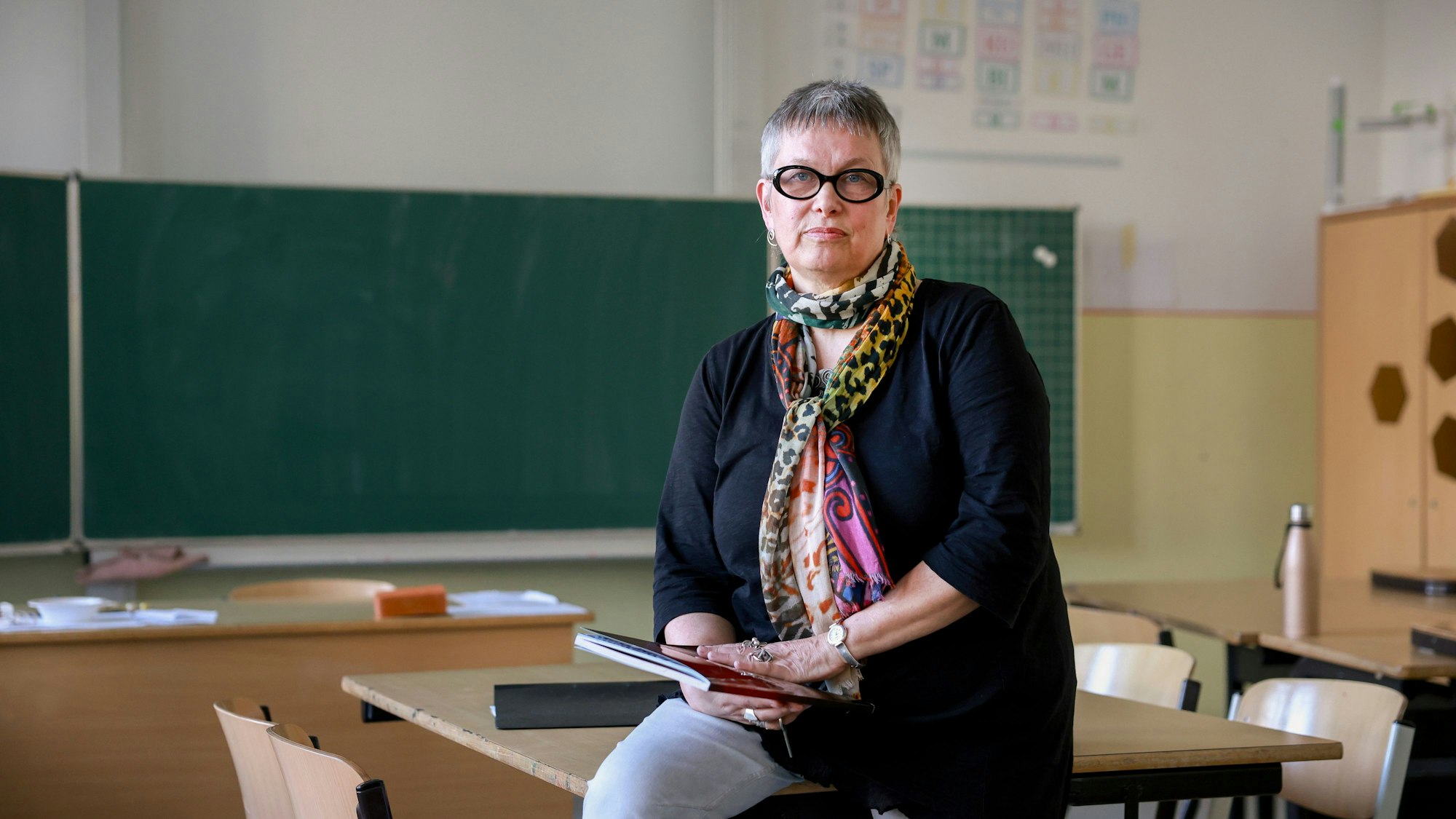 Silke Micheel sitzt halb auf einem Tisch in einem Klassenraum. In den Händen hält sie ein Heft.