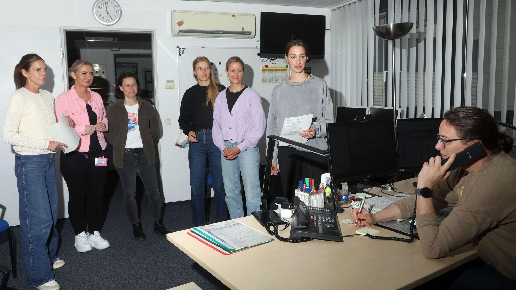 30.03.2025, Köln: Doro (v.l.), Kristin, Simone, Milena , Imke und Carina stehen im Büro der Wachdienstleiterin Vanessa, die an einem Schreibtisch telefoniert.