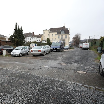 Parkende Autos auf einer Schotterfläche, links an der Seite ein Wohnhaus.