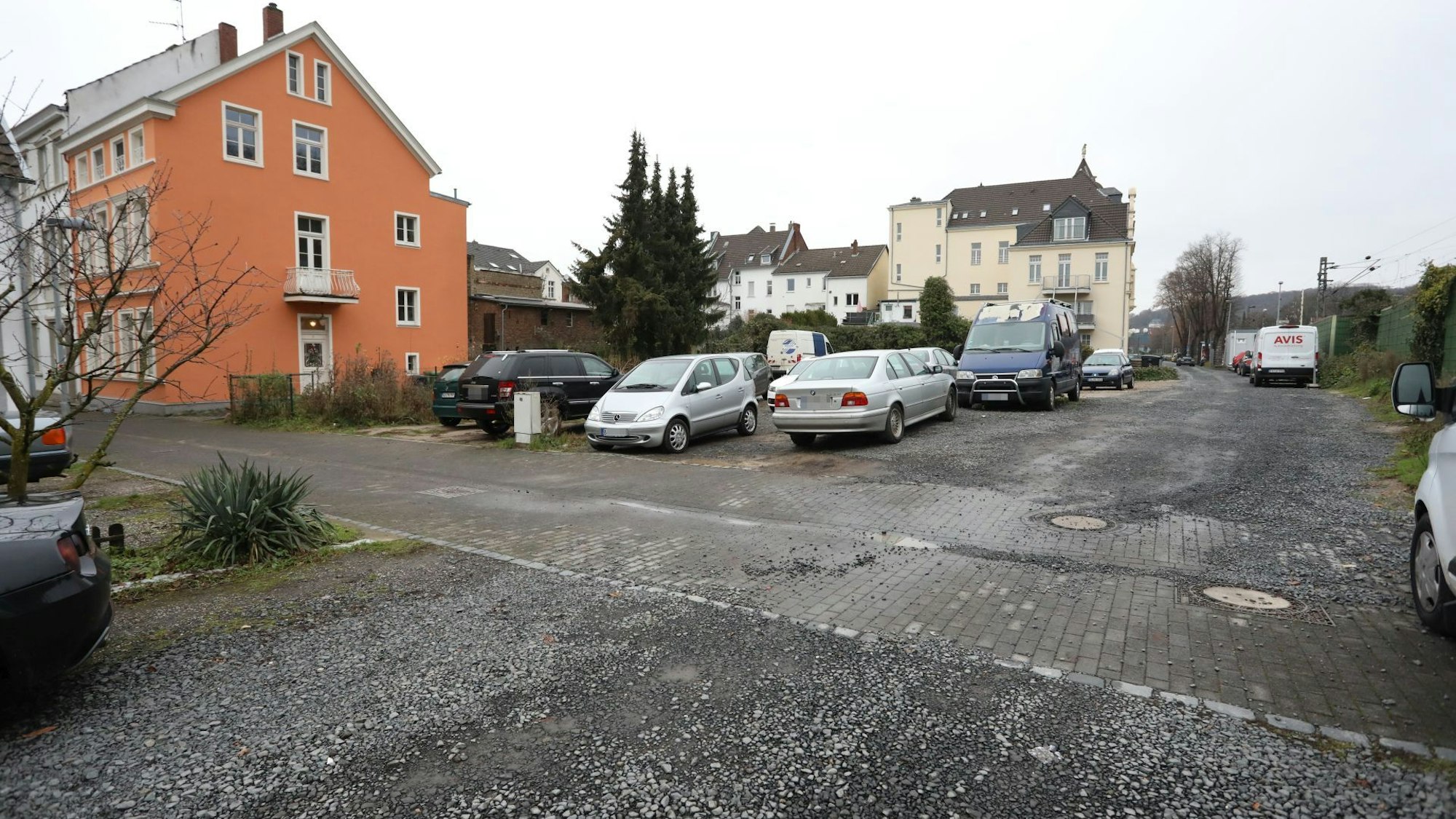 Parkende Autos auf einer Schotterfläche, links an der Seite ein Wohnhaus.