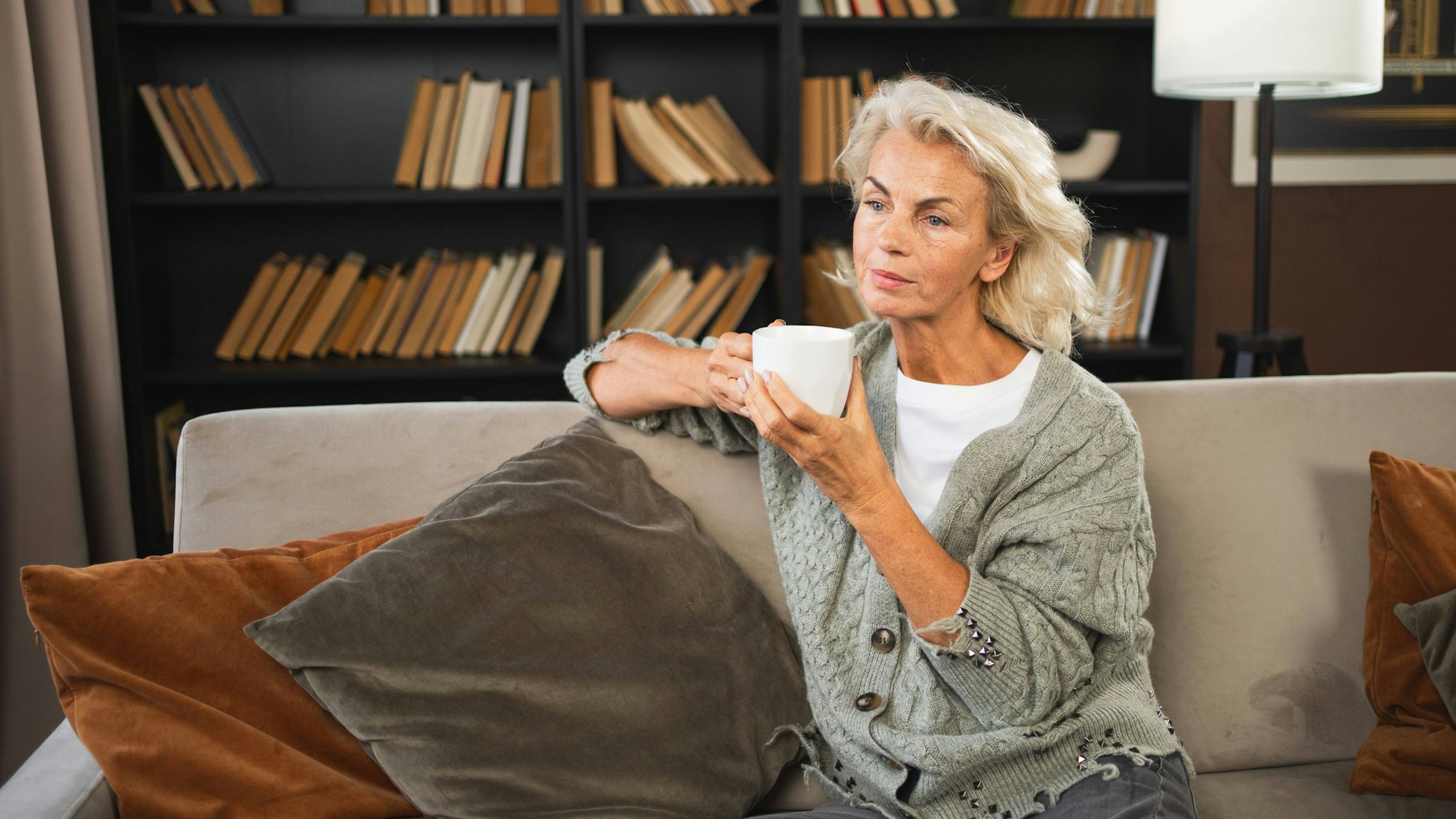 Eine Frau sitzt auf einem Sofa, hat eine Tasse in der Hand und starrt ins Leere.
