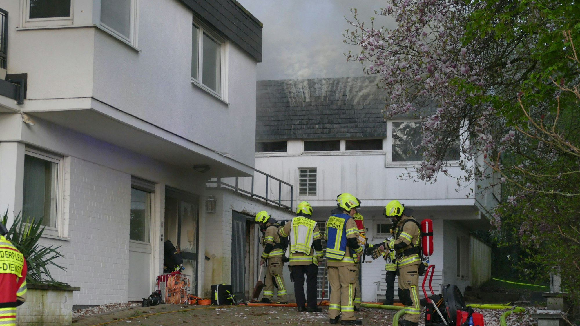 Feuerwehrleute stehen vor den beiden Gebäudeteilen des Hauses in Sankt Augustin-Birlinghoven, dichter Rauch quillt aus dem Dach.
