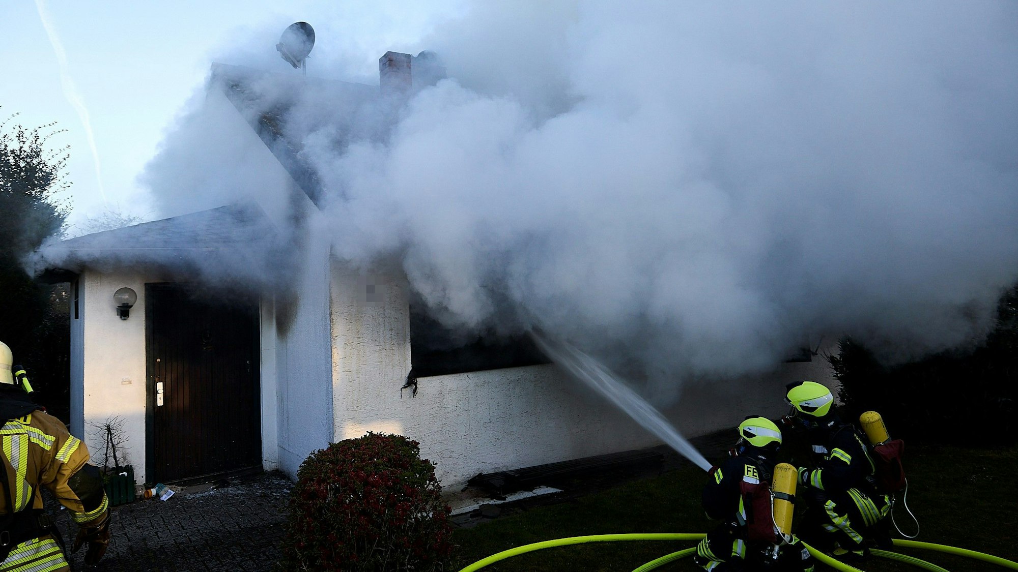 Feuerwehrleute geben durch ein Fenster Wasser in ein Wohnhaus ab, aus dem dichter Rauch austritt.
