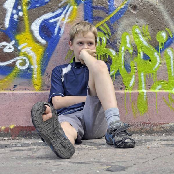 Ein trauriger, lustloser Junge sitzt vor einer Graffiti-Mauer, in einer seiner Schuhsohlen klafft ein großes Loch.