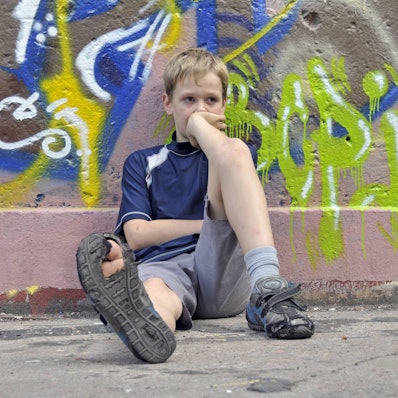 Ein trauriger, lustloser Junge sitzt vor einer Graffiti-Mauer, in einer seiner Schuhsohlen klafft ein großes Loch.