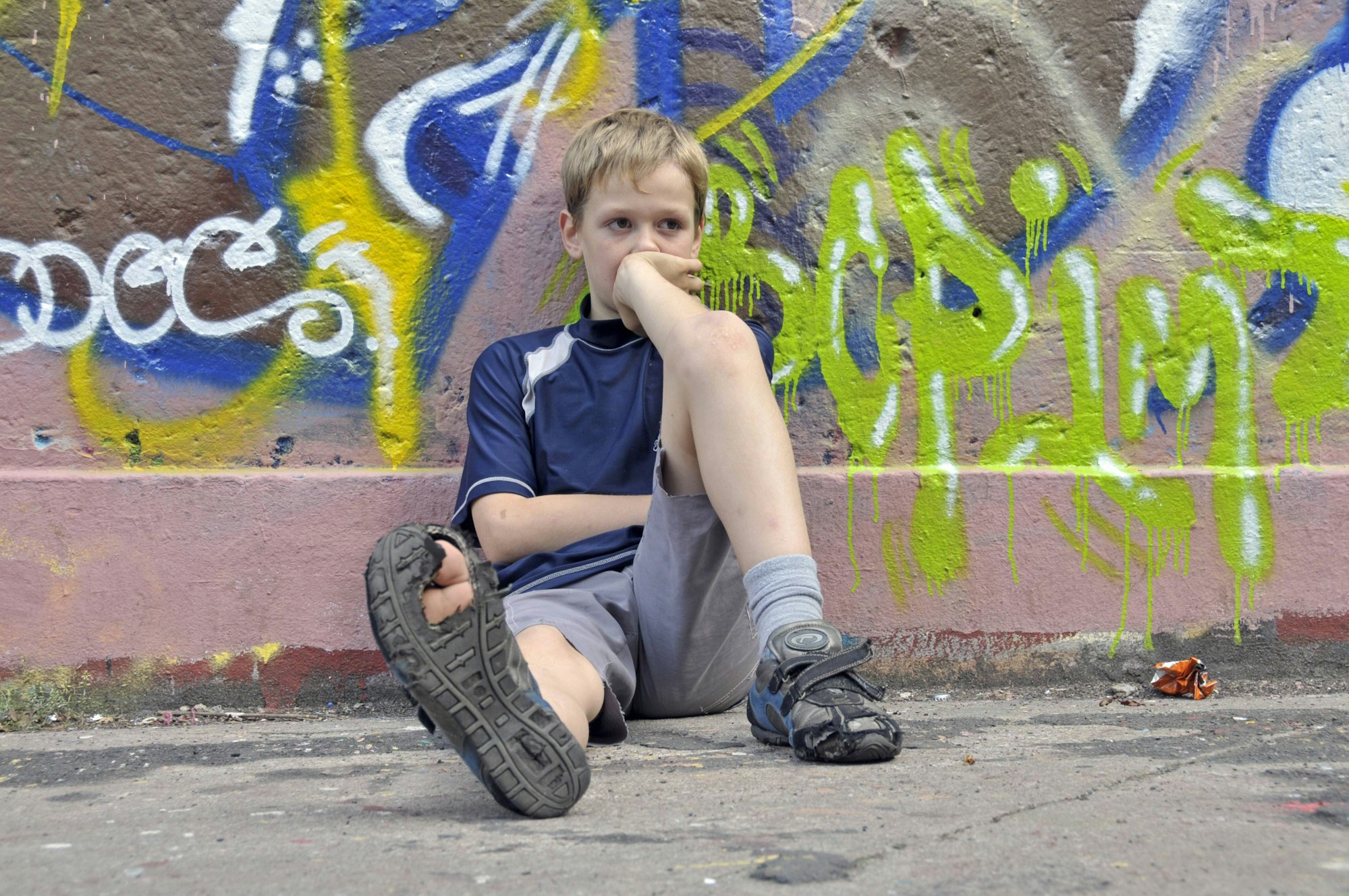Ein trauriger, lustloser Junge sitzt vor einer Graffiti-Mauer, in einer seiner Schuhsohlen klafft ein großes Loch.