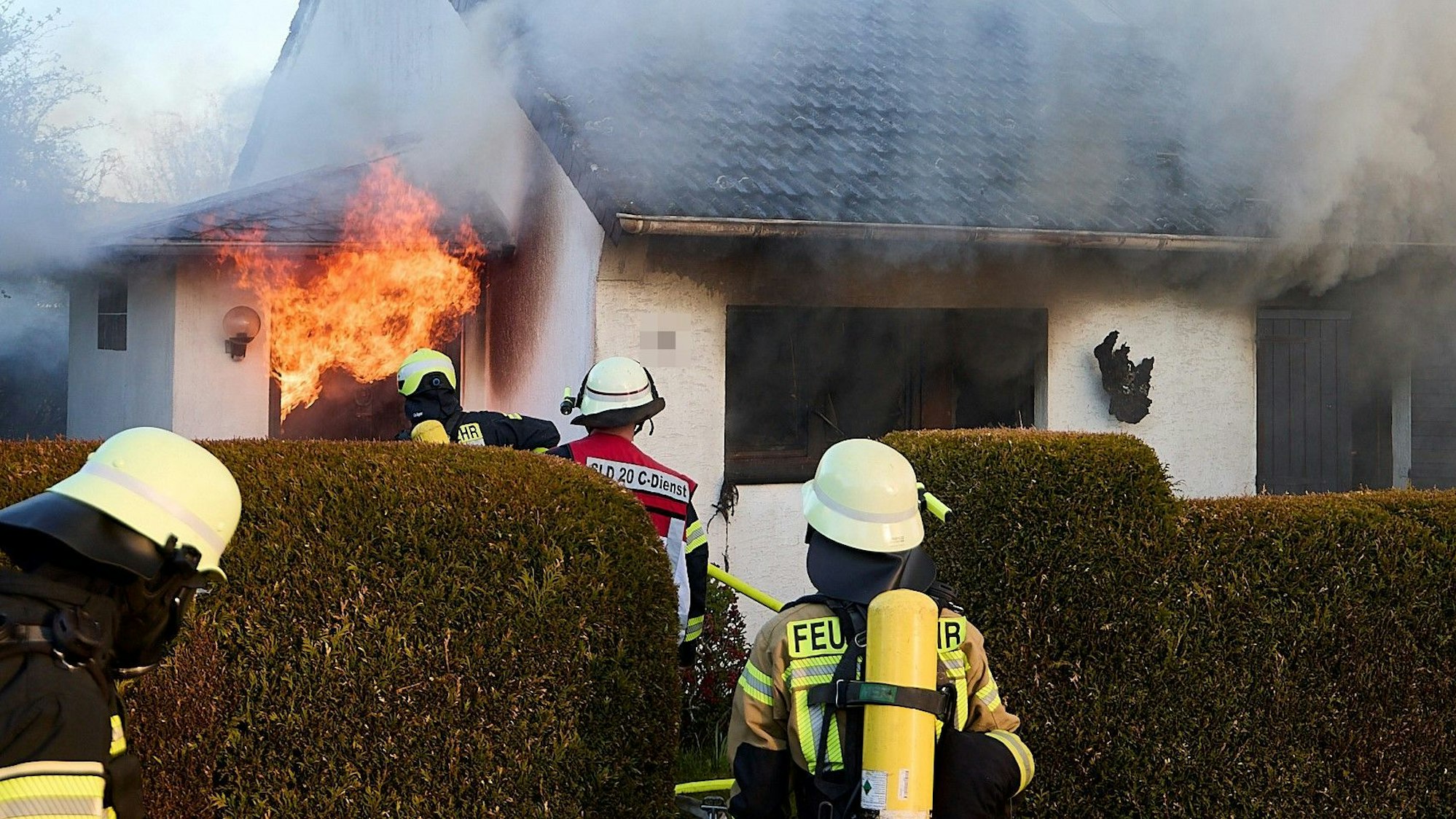 Feuerwehrleute gehen in Richtung eines Wohnhauses, aus dem Flammen schlagen. Rauchwolken steigen aus dem Gebäude auf.