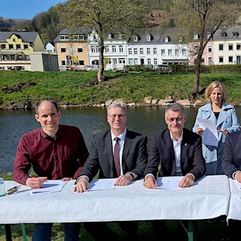 Fünf Männer und eine Frau sitzen an einem Tisch vor einem Fluss. Hinter ihnen stehen ein Mann und eine Frau. Im Hintergrund stehen Häuser und Bäume.