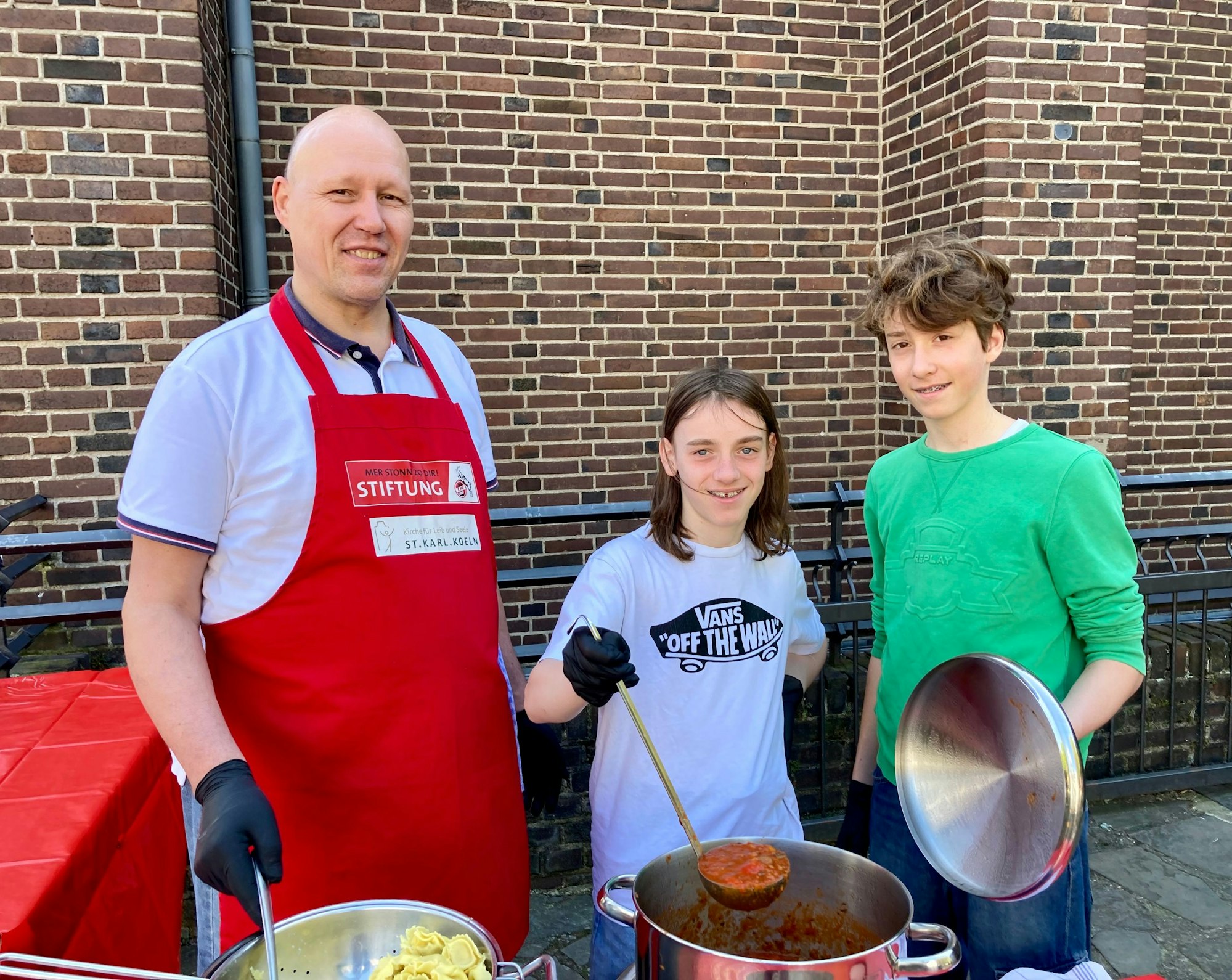 Zwei Teenager und ein älterer Mann stehen hinter großen Essenstöpfen und haben Suppenkellen in der Hand.
