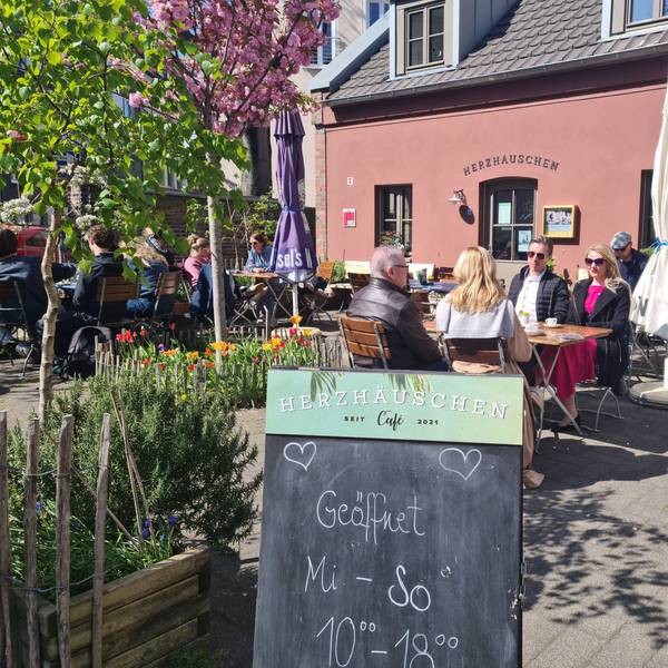 Auf einer Café-Terrasse vor einem Terrakotta-roten Gebäude sitzen viele Menschen an Tischen. Im Vordergrund steht eine Tafel, auf der steht: Geöffnet Mi.–So., 10–18 Uhr.