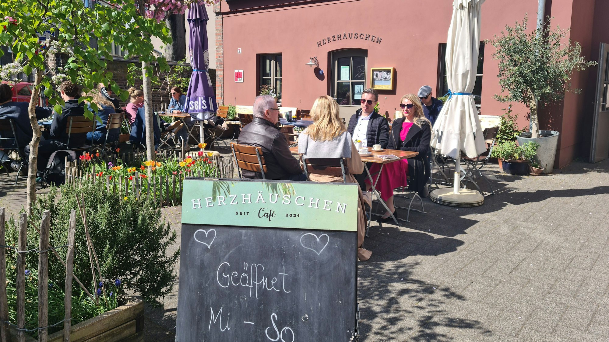Auf einer Café-Terrasse vor einem Terrakotta-roten Gebäude sitzen viele Menschen an Tischen. Im Vordergrund steht eine Tafel, auf der steht: Geöffnet Mi.–So., 10–18 Uhr.