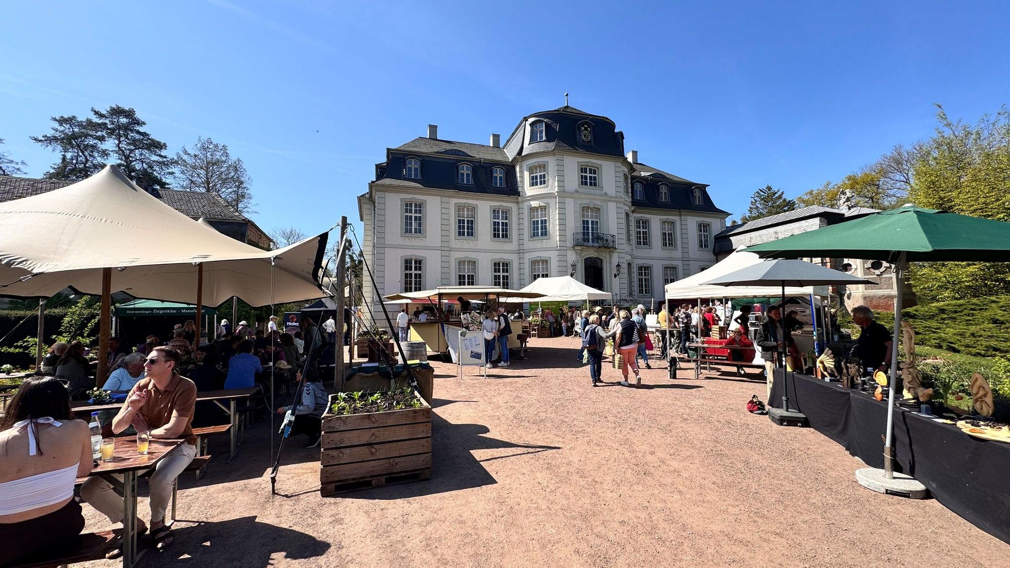 Von Sonnenschirmen geschützte Stände, im Hintergrund ein Schloss mit einer hellen Fassade.