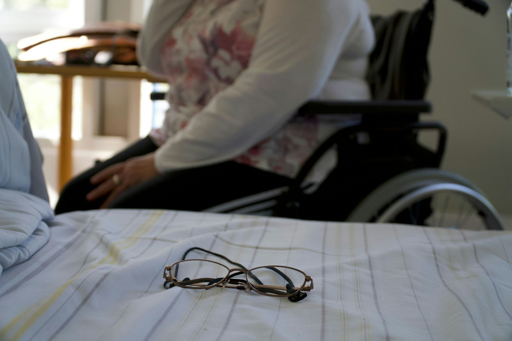 Oya Kar sitzt im Rollstuhl vor ihrem Bett, auf dem eine Brille liegt
