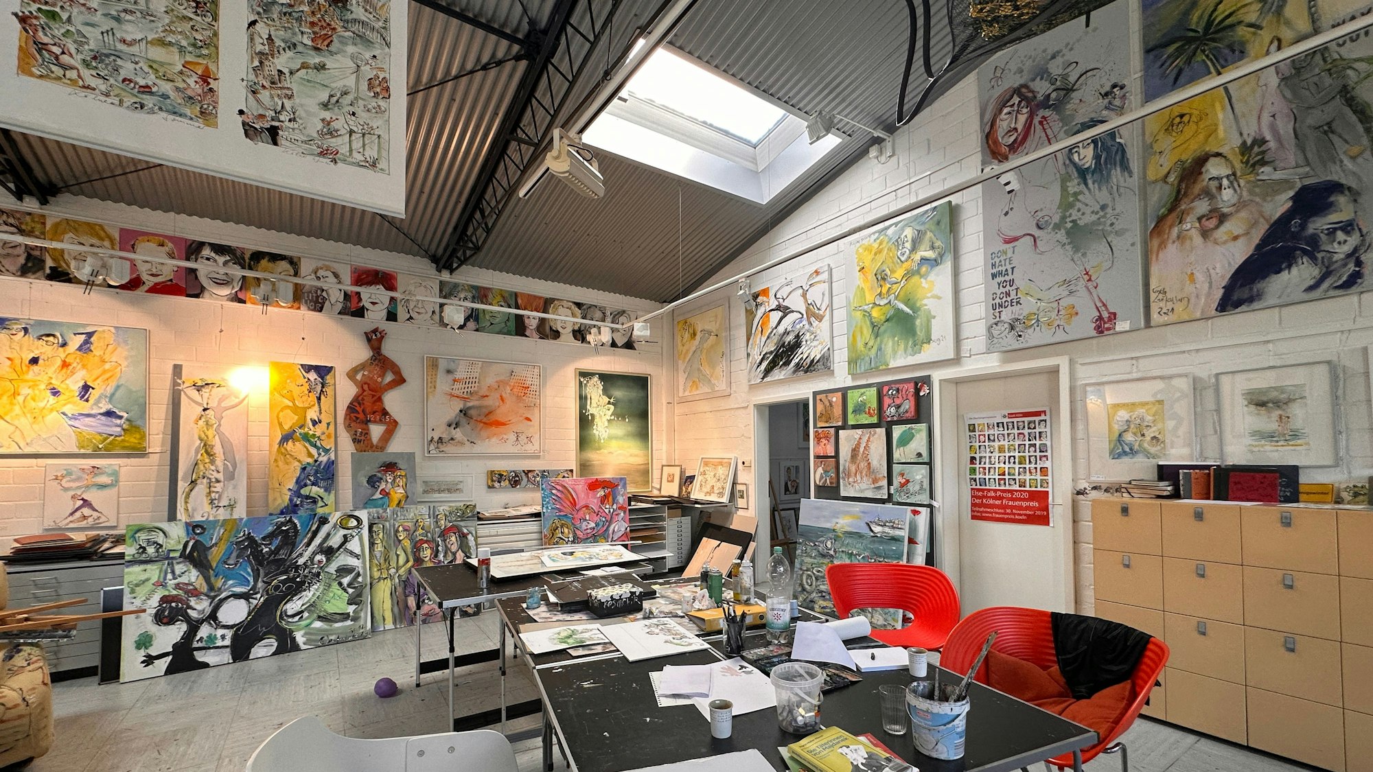 Blick in einen hohen Raum, an dem ein Tisch in der Mitte steht, an den Wänden lehnen und hängen zahlreiche Bilder.