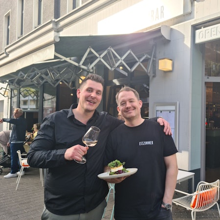 Zwei junge Männer in schwarzen Oberteilen stehen vor der geöffneten Tür eines Ecklokals.