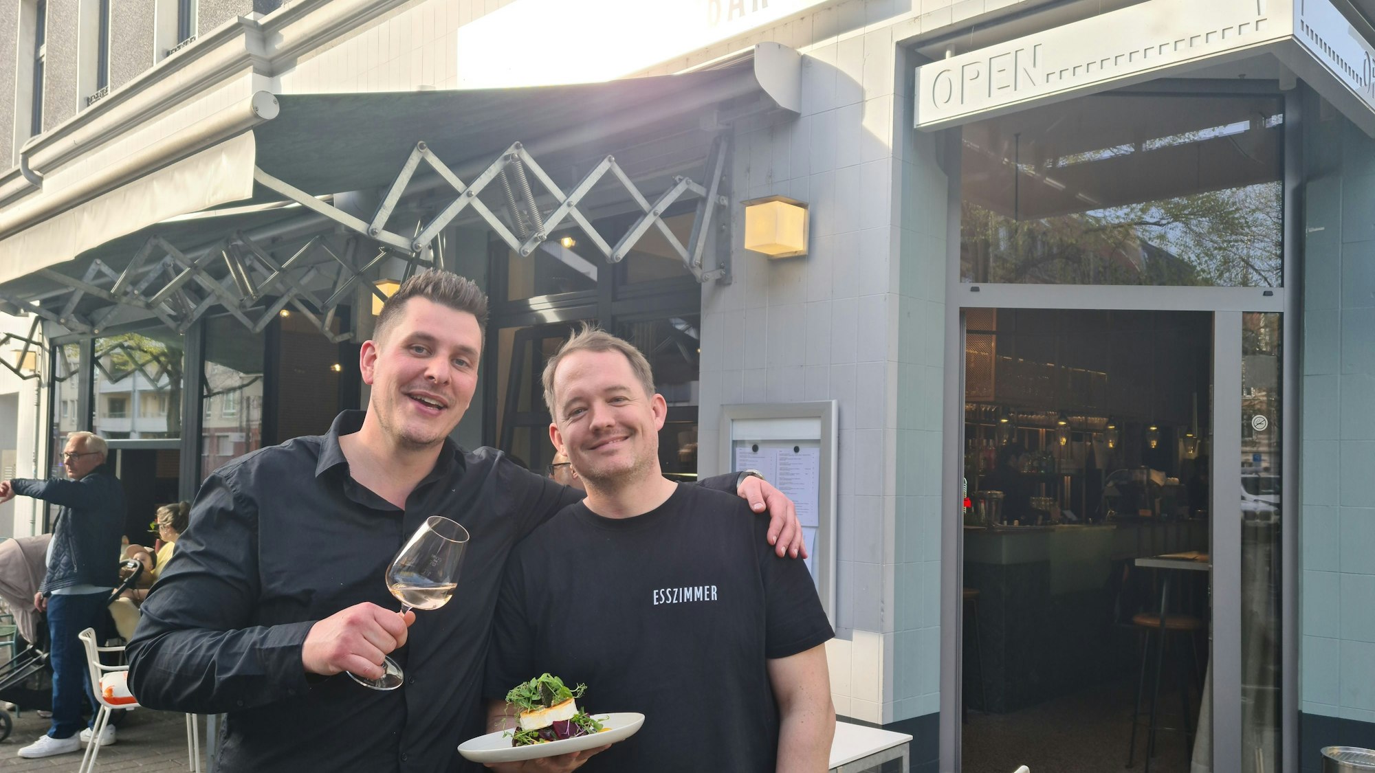 Zwei junge Männer in schwarzen Oberteilen stehen vor der geöffneten Tür eines Ecklokals.