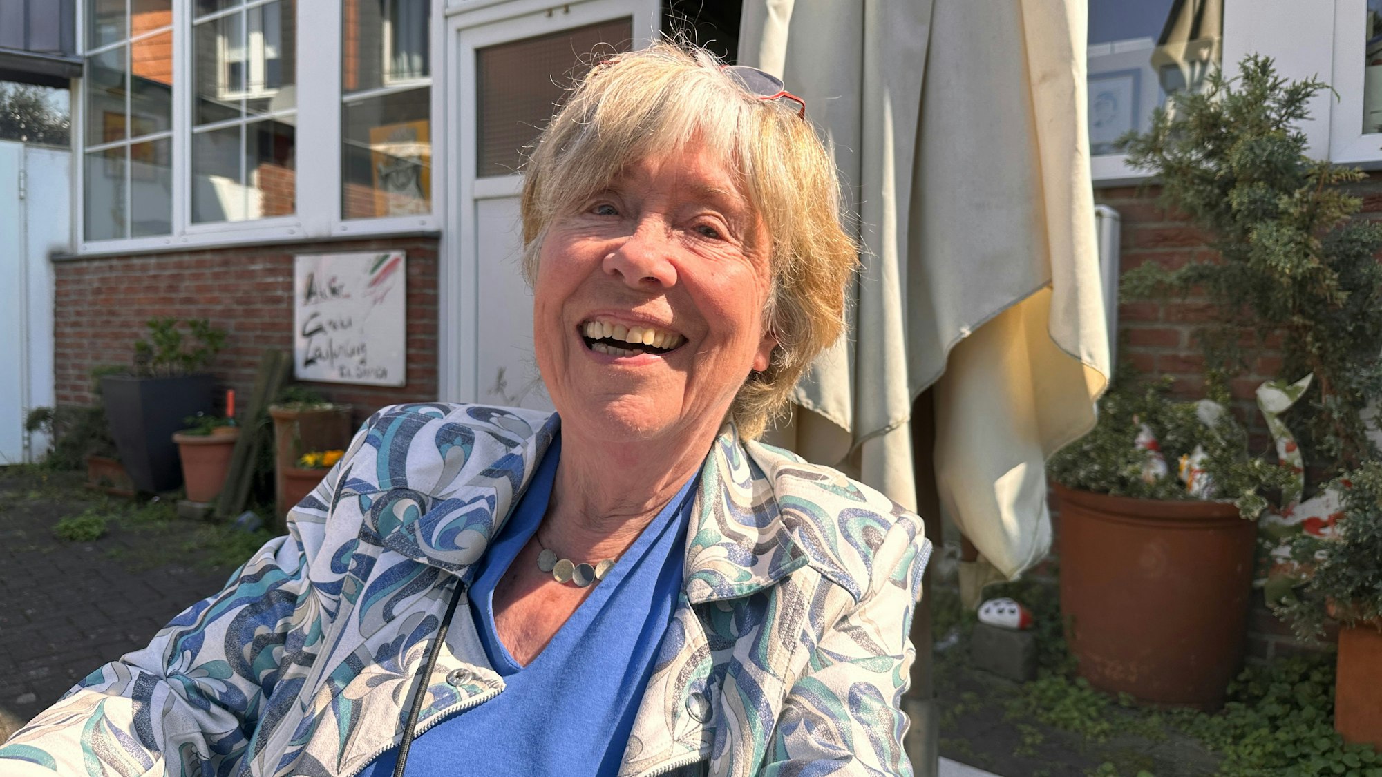 Eine ältere Dame, die vor einem Gebäude auf einer Terrasse sitzt, lacht herzhaft in die Kamera.