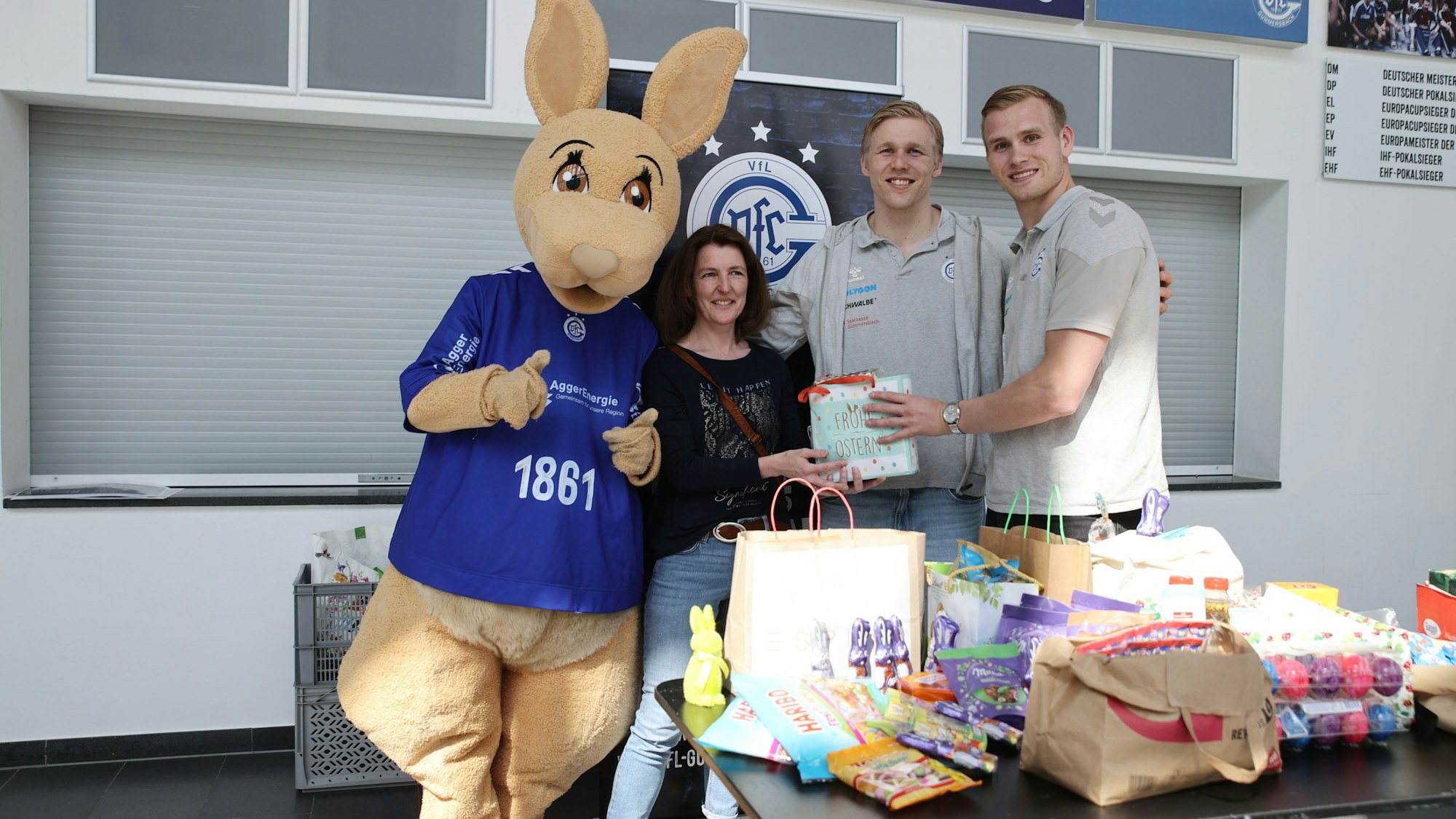 Zwei VfL-Handballer und Maskottchen Gummi nehmen Geschenke für Ostern entgegen. Eine Frau reicht ihnen eine Tüte.