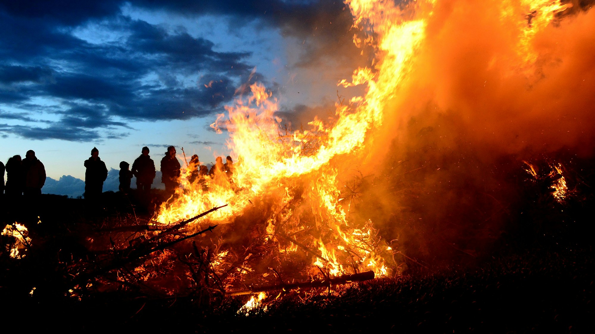 ARCHIV - 15.04.2017, Niedersachsen, Wartjenstedt: Zuschauer stehen bei einem traditionellen Osterfeuer. Wegen der anhaltenden Trockenheit diskutieren zahlreiche Kommunen in Nordrhein-Westfalen, ob die traditionellen Osterfeuer in diesem Jahr stattfinden können. In mehreren Kreisen etwa im Sauerland sind die Ordnungsämter im Austausch mit der Feuerwehr. (zu dpa: «Trockenheit könnte zu Absagen bei Osterfeuern führen») Foto: Maurizio Gambarini/dpa +++ dpa-Bildfunk +++
