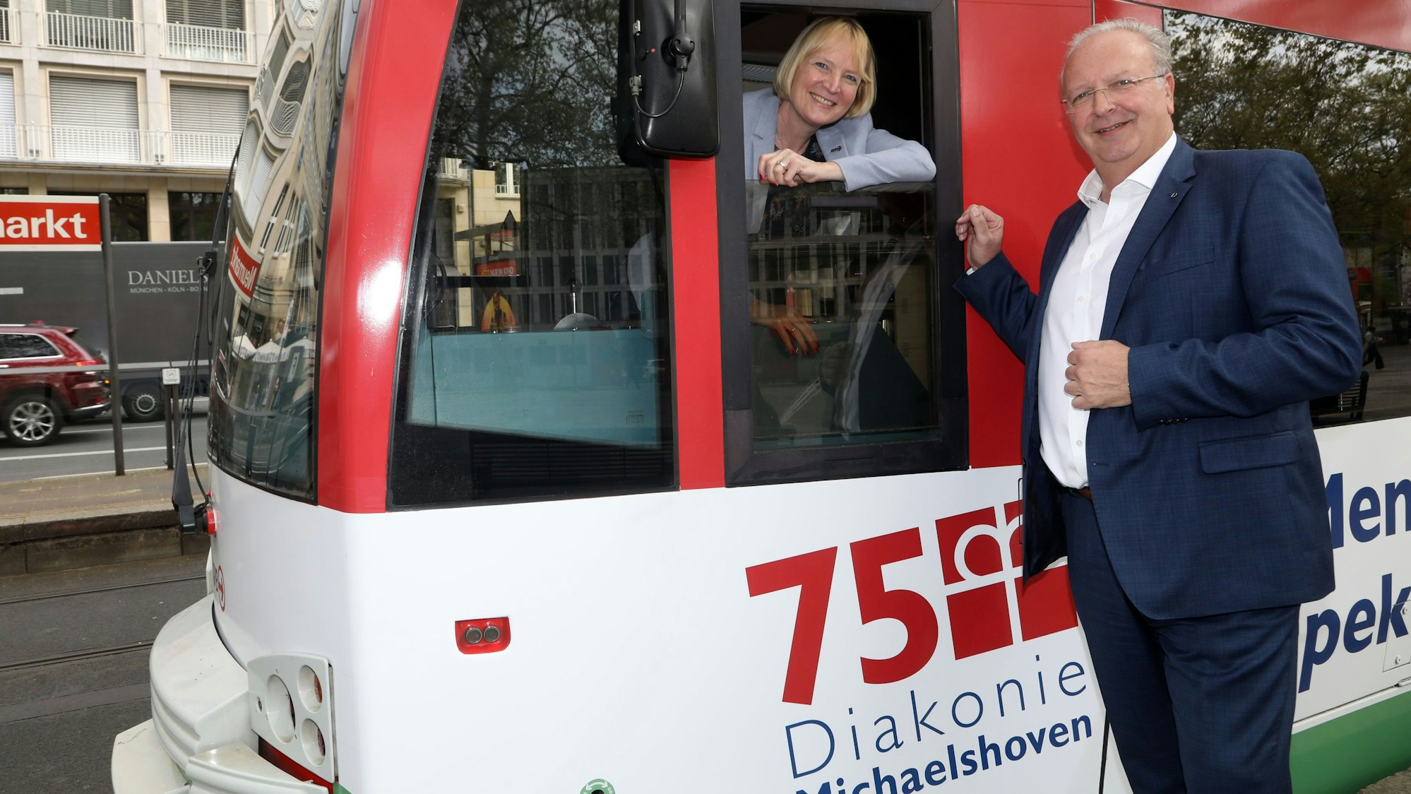 Zu sehen ist auf der linken Seite Stefanie Haaks, Vorstandsvorsitzende der KVB, die aus dem Fenster der Fahrerkabine einer Bahn der KVB herausschaut. Auf der rechten Seite ist Uwe Ufer zu sehen, der neben der Bahn steht.