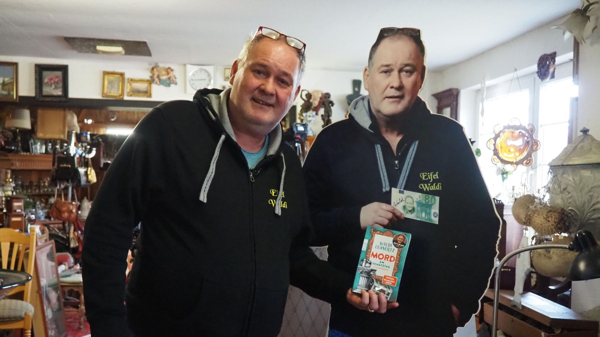 Ein Mann im Pullover und mit einer Brille, die auf seinem Kopf aufliegt, steht neben einem Pappaufsteller von sich und hält ein Buch in die Kamera.