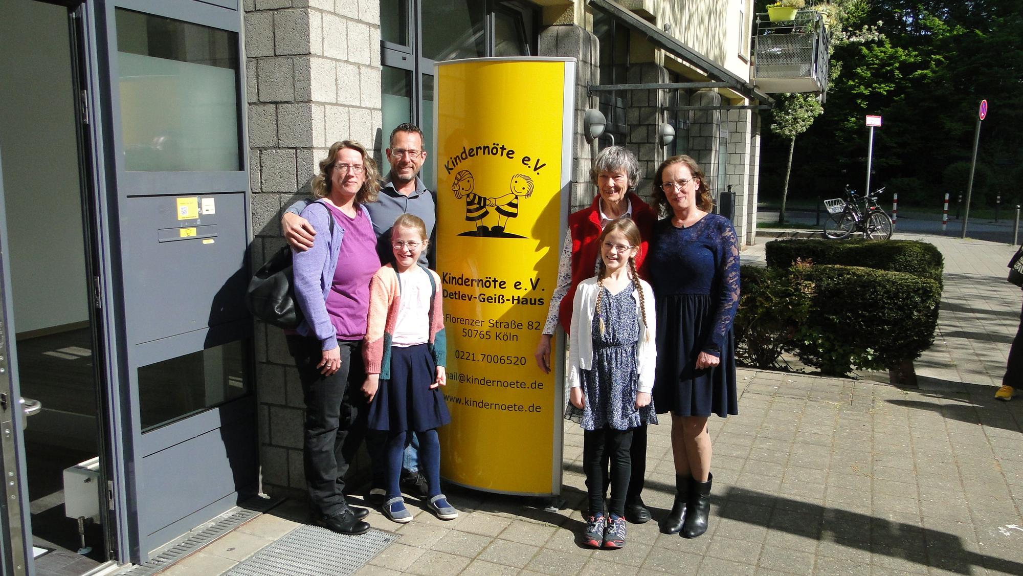 Vier Erwachsene und zwei Kinder stehen vor einem Hauseingang links und rechtrs von einer gelben Säule mit den Informationen zum neuen Detlev-Geiß-Haus.