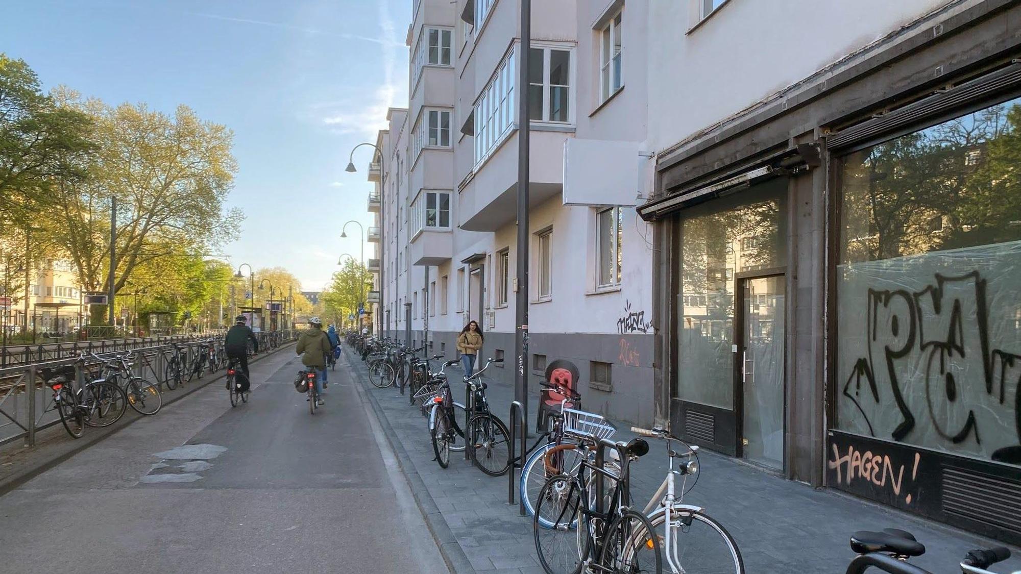 Fahrradstraße auf der Zülpicher Straße, auf der Straße fahren zwei Fahrräder, auf dem Gehweg geht eine Frau. Die neu gebauten Fahrradständer am Rand des Gehwegs sind größtenteils belegt.