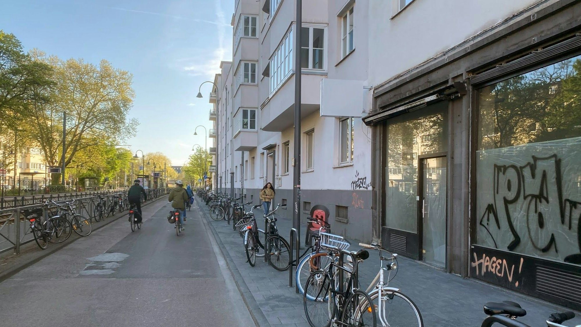Fahrradstraße auf der Zülpicher Straße, auf der Straße fahren zwei Fahrräder, auf dem Gehweg geht eine Frau. Die neu gebauten Fahrradständer am Rand des Gehwegs sind größtenteils belegt.