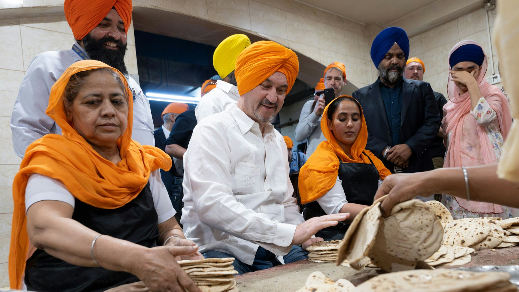 13.04.2025, Indien, Neu Delhi: Markus Söder, (CSU, M) Ministerpräsident von Bayern, besucht die Großküche des Sikh-Tempel «Gurudwara Bangla Sahib» mit einem orangenen Turban auf dem Kopf. Dabei lässt er sich zeigen, wie traditionelles Fladenbrot gemacht wird. Söder absolviert eine mehrtägigen Indienreise. Foto: Peter Kneffel/dpa +++ dpa-Bildfunk +++