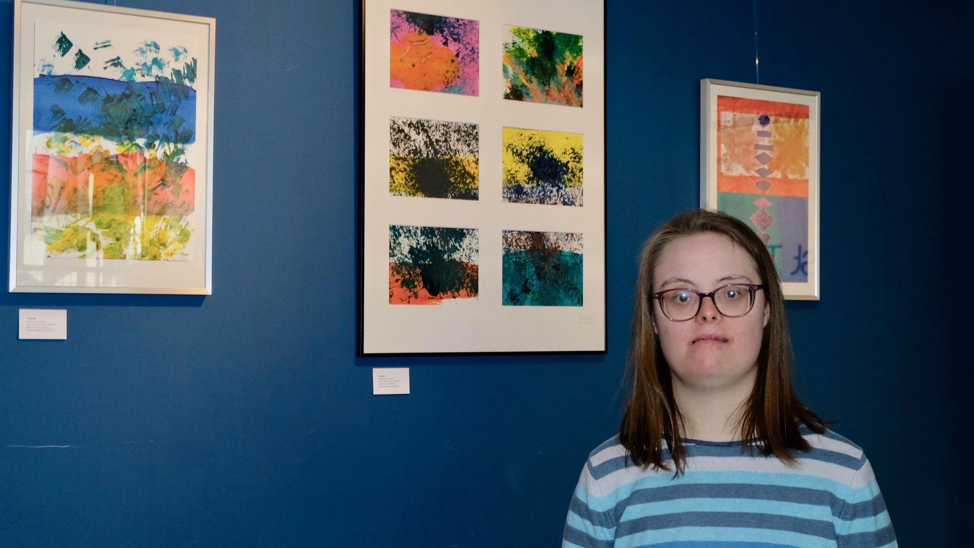 Eine junge Frau mit schulterlangem Haar und Brille steht vor einer blauen Wand, an der drei abstrakte Bilder in Pastellfarben hängen.