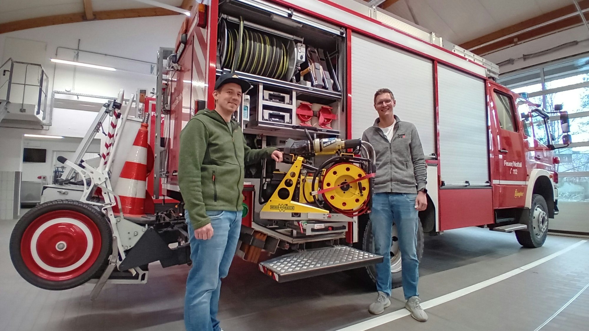 Zwei Feuerwehrleute stehen in einer Halle an einem Einsatzwagen der Feuerwehr.