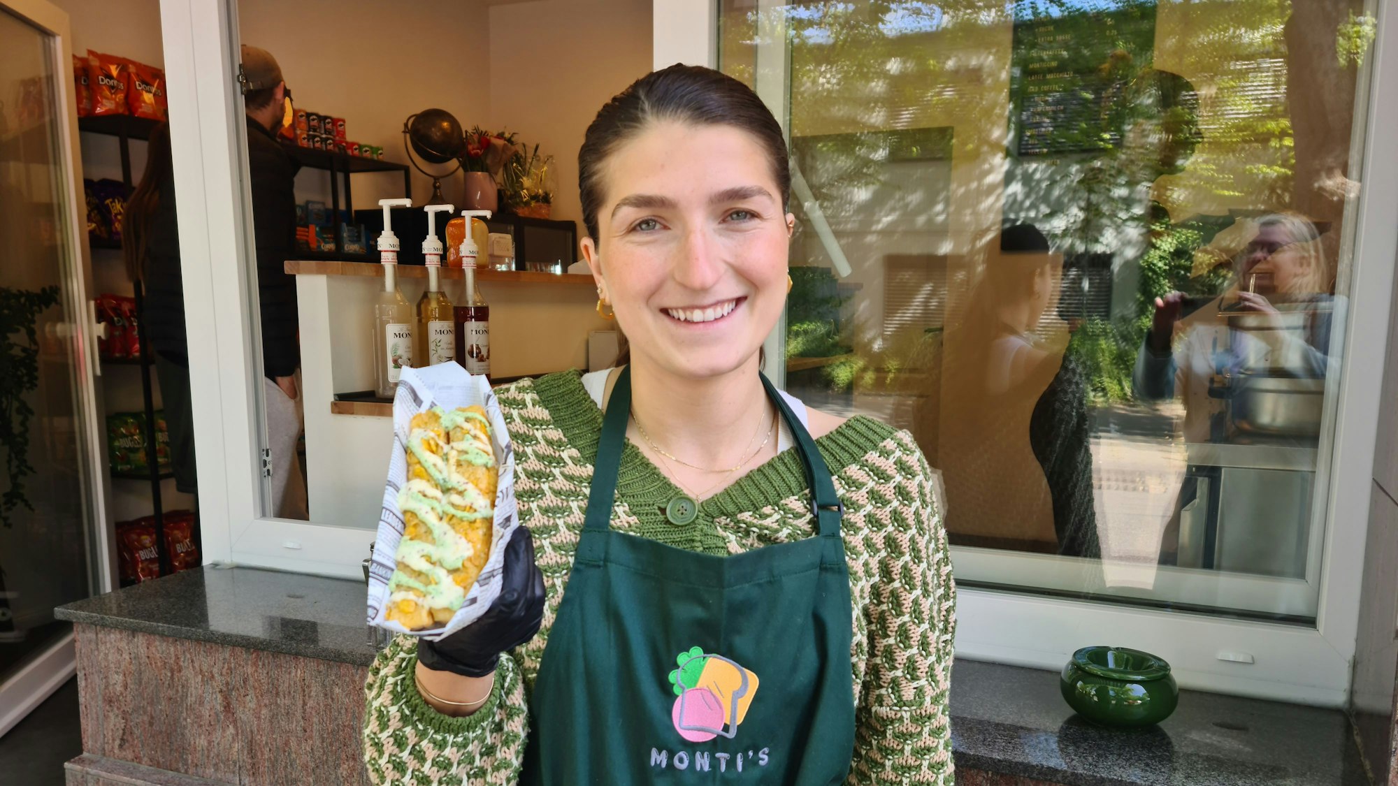 Eine junge Frau mit Pferdeschwanz und grüner Schürze präsentiert ein French Toast. Sie steht vor dem Fenster, das halb geöffnet ist, und der offenen Tür eines Kiosks.