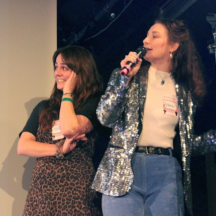 Ivana redet auf einer Bühne in ein Mikrofon und schaut dabei auf eine Leinwand. Neben ihr steht Teresa, die etwas peinlich berührt ebenfalls zur Leinwand schaut.