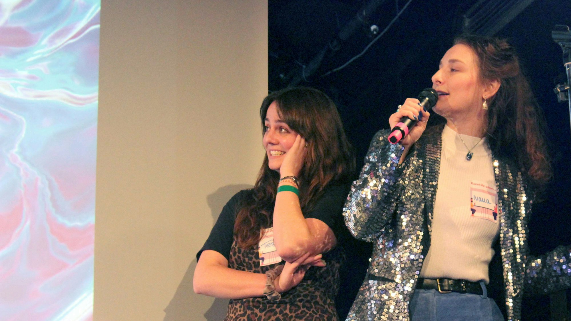 Ivana redet auf einer Bühne in ein Mikrofon und schaut dabei auf eine Leinwand. Neben ihr steht Teresa, die etwas peinlich berührt ebenfalls zur Leinwand schaut.