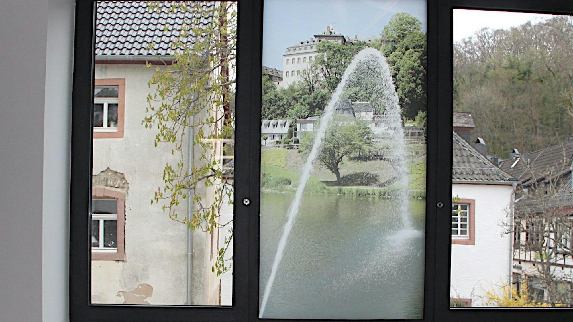 Im mittleren Feld eines dreiteiligen Fensters ist eine transparente Folie aufgeklebt, die eine Wasserfontäne und die Burg Blankenheim zeigt.