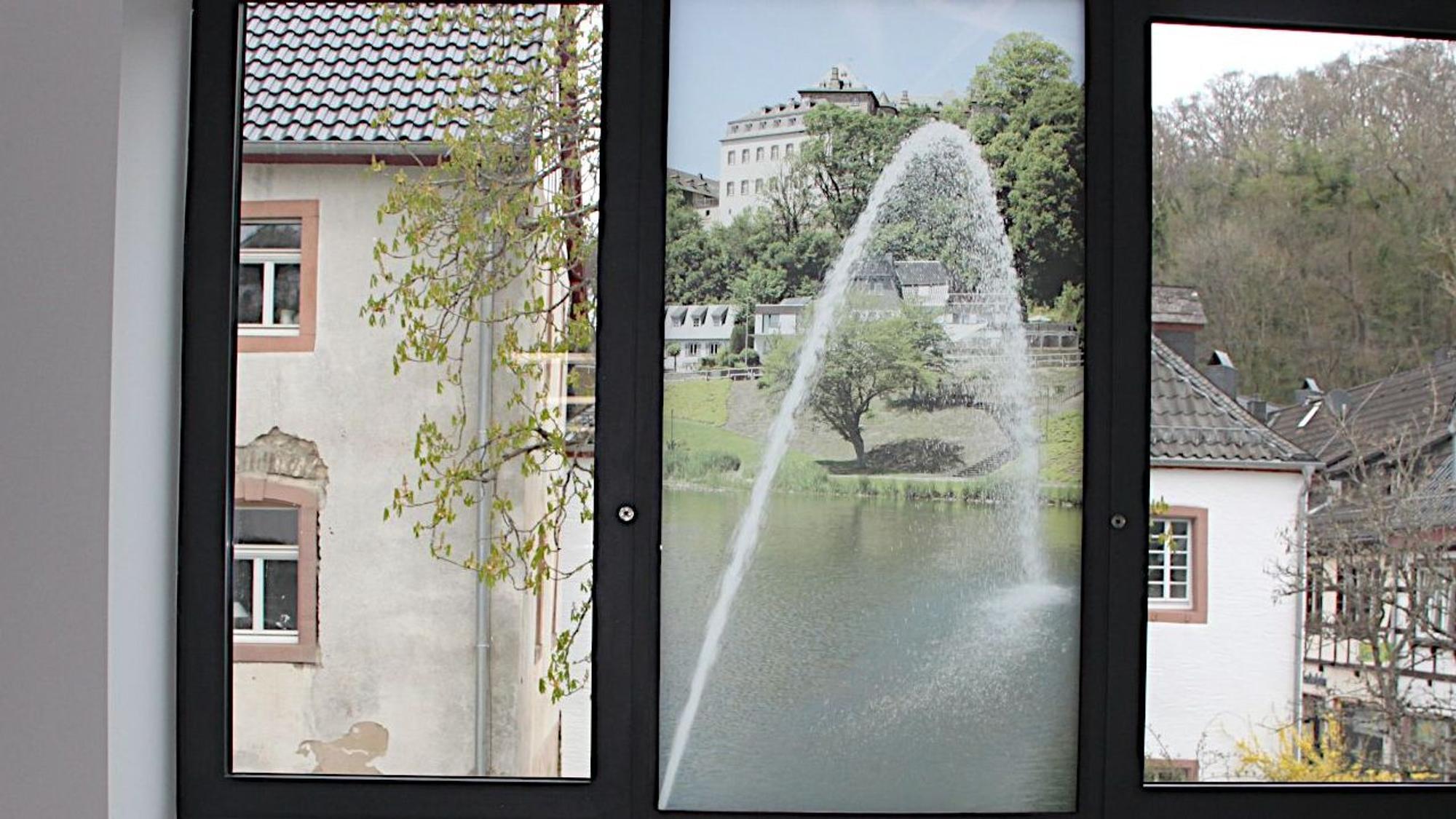 Im mittleren Feld eines dreiteiligen Fensters ist eine transparente Folie aufgeklebt, die eine Wasserfontäne und die Burg Blankenheim zeigt.