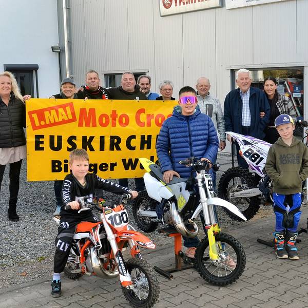 Fünf Kinder und Jugendliche stehen an Motocross-Maschinen, dahinter einige Erwachsene, die mit einem Plakat auf den Wettbewerb am 1. Mai im Billiger Wald hinweisen.