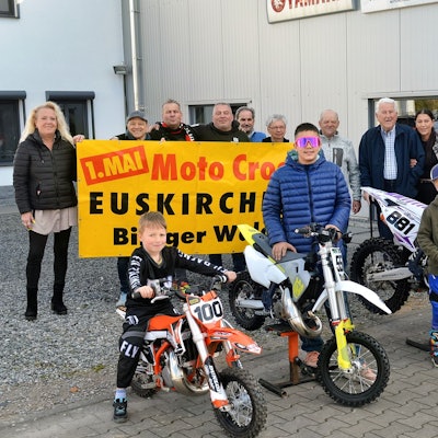 Fünf Kinder und Jugendliche stehen an Motocross-Maschinen, dahinter einige Erwachsene, die mit einem Plakat auf den Wettbewerb am 1. Mai im Billiger Wald hinweisen.