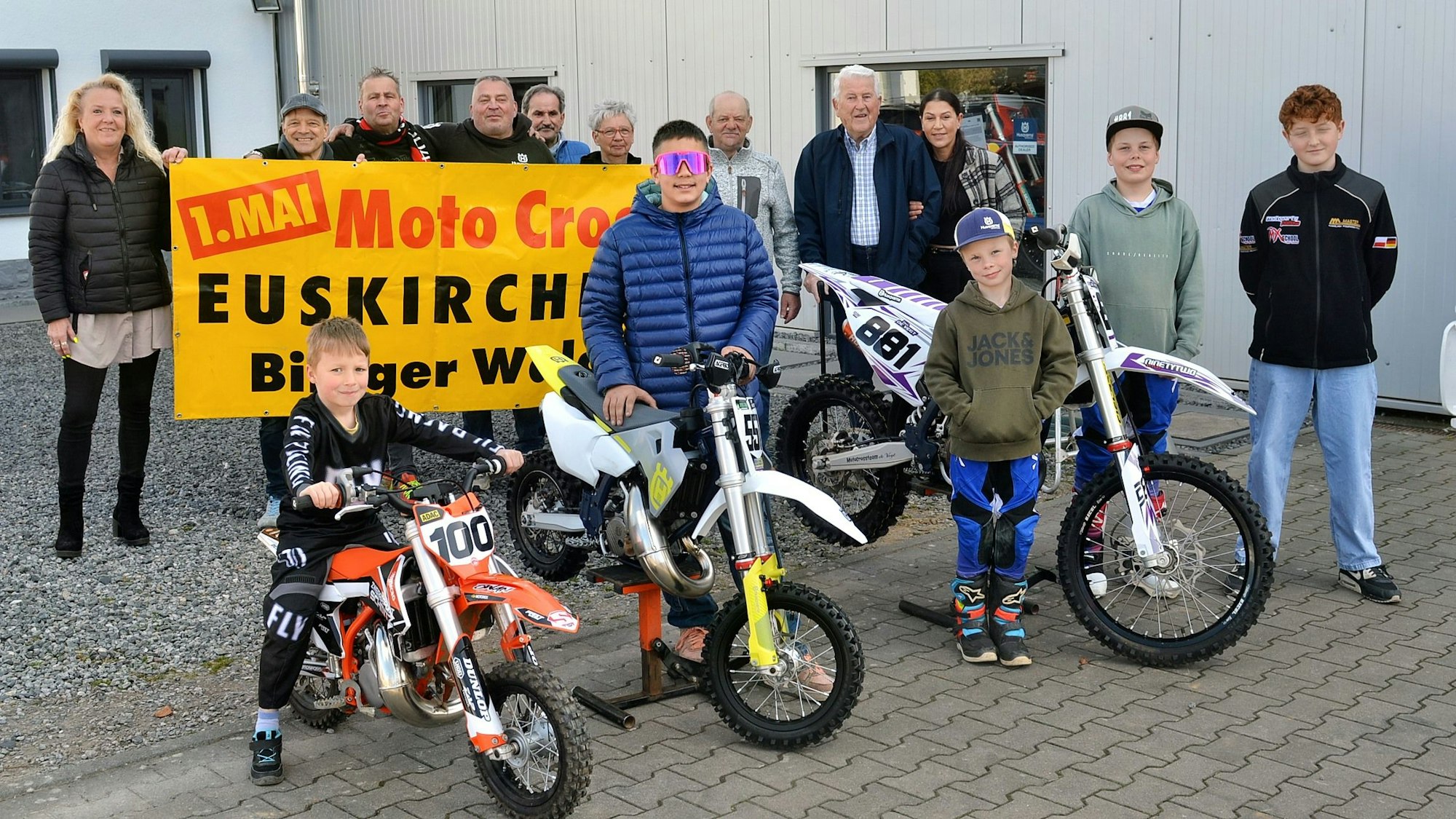 Fünf Kinder und Jugendliche stehen an Motocross-Maschinen, dahinter einige Erwachsene, die mit einem Plakat auf den Wettbewerb am 1. Mai im Billiger Wald hinweisen.