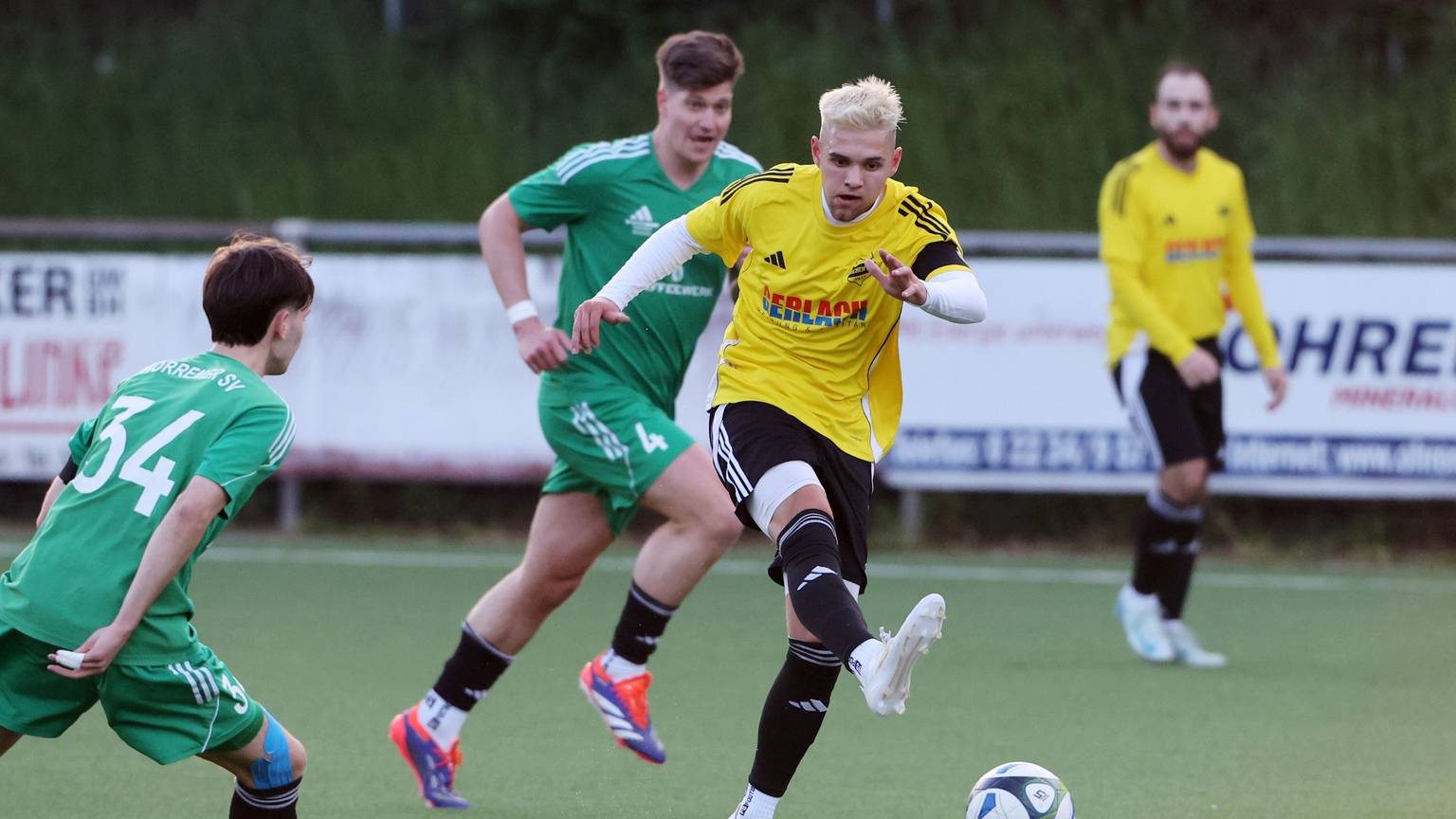 Das Foto zeigt einen Spieler des VfR Bachem im gelben Trikot, der von zwei Horremern in grünen Trikots am Abspiel nicht gehindert werden kann.