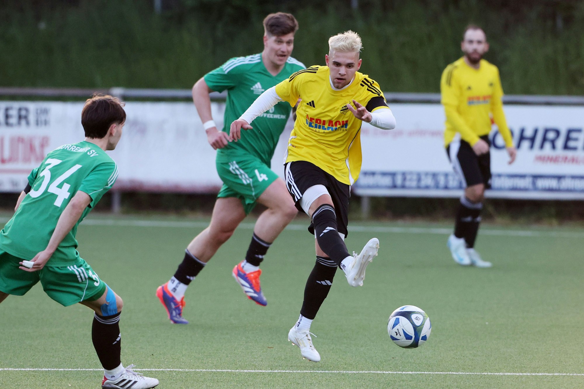 Das Foto zeigt einen Spieler des VfR Bachem im gelben Trikot, der von zwei Horremern in grünen Trikots am Abspiel nicht gehindert werden kann.
