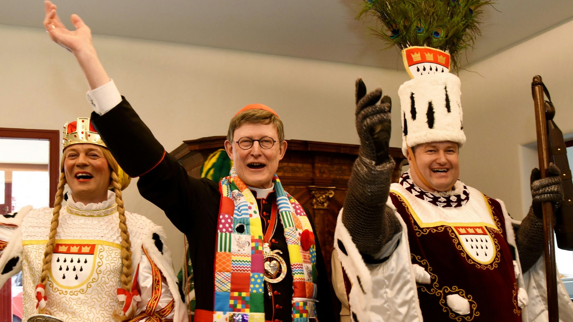 Kardinal Rainer Woelki macht zwischen zwei Mitgleidern des Kölner Dreigestirns von 2020 eine ausladende Geste. Er trägt einen bunten Schal und einen Karnevalsorden.