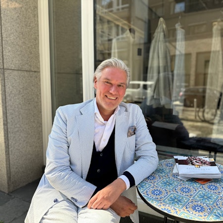 Ein Mann mit einem blau-weiß-gestreiften Anzug sitzt an einem Tisch, der mit Blumen-Fliesen gemustert ist.