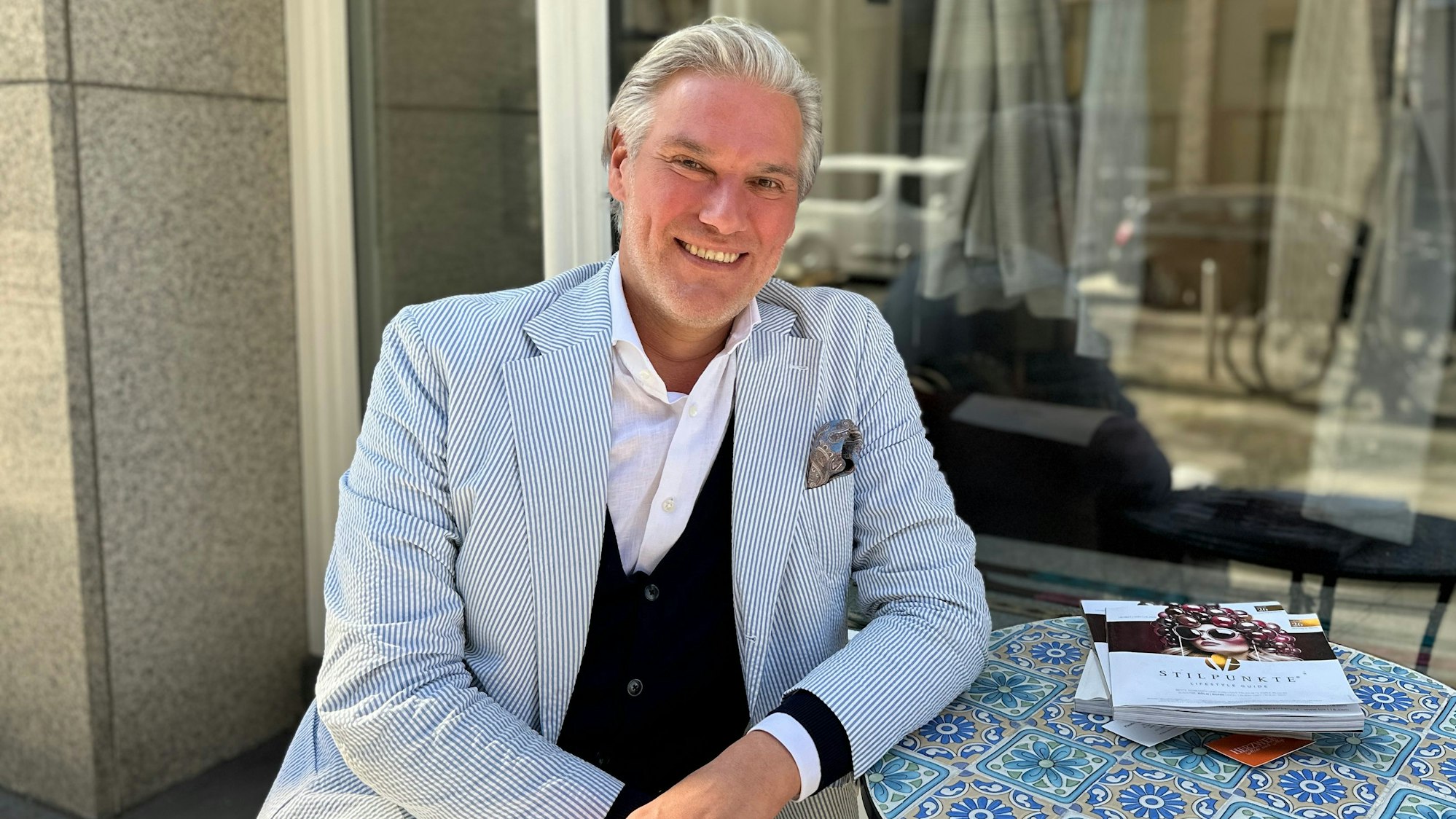 Ein Mann mit einem blau-weiß-gestreiften Anzug sitzt an einem Tisch, der mit Blumen-Fliesen gemustert ist.