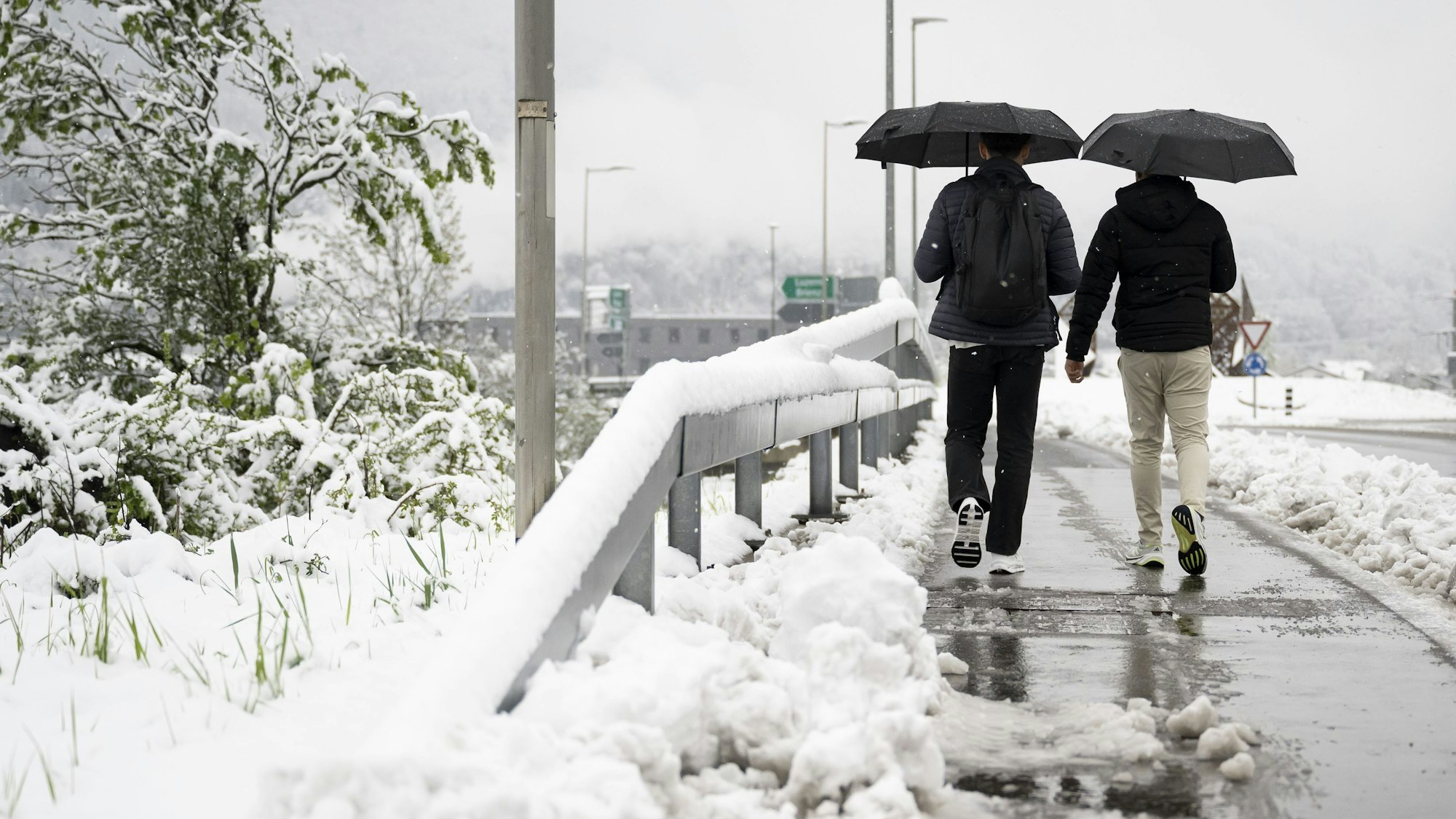 Zwei Personen gehen auf einem teilweise vom Schnee geräumten Bürgersteig auf einer Brücke über die Autobahn zwischen Interlaken-West und Interlaken-Ost in Wilderswil im Berner Oberland.