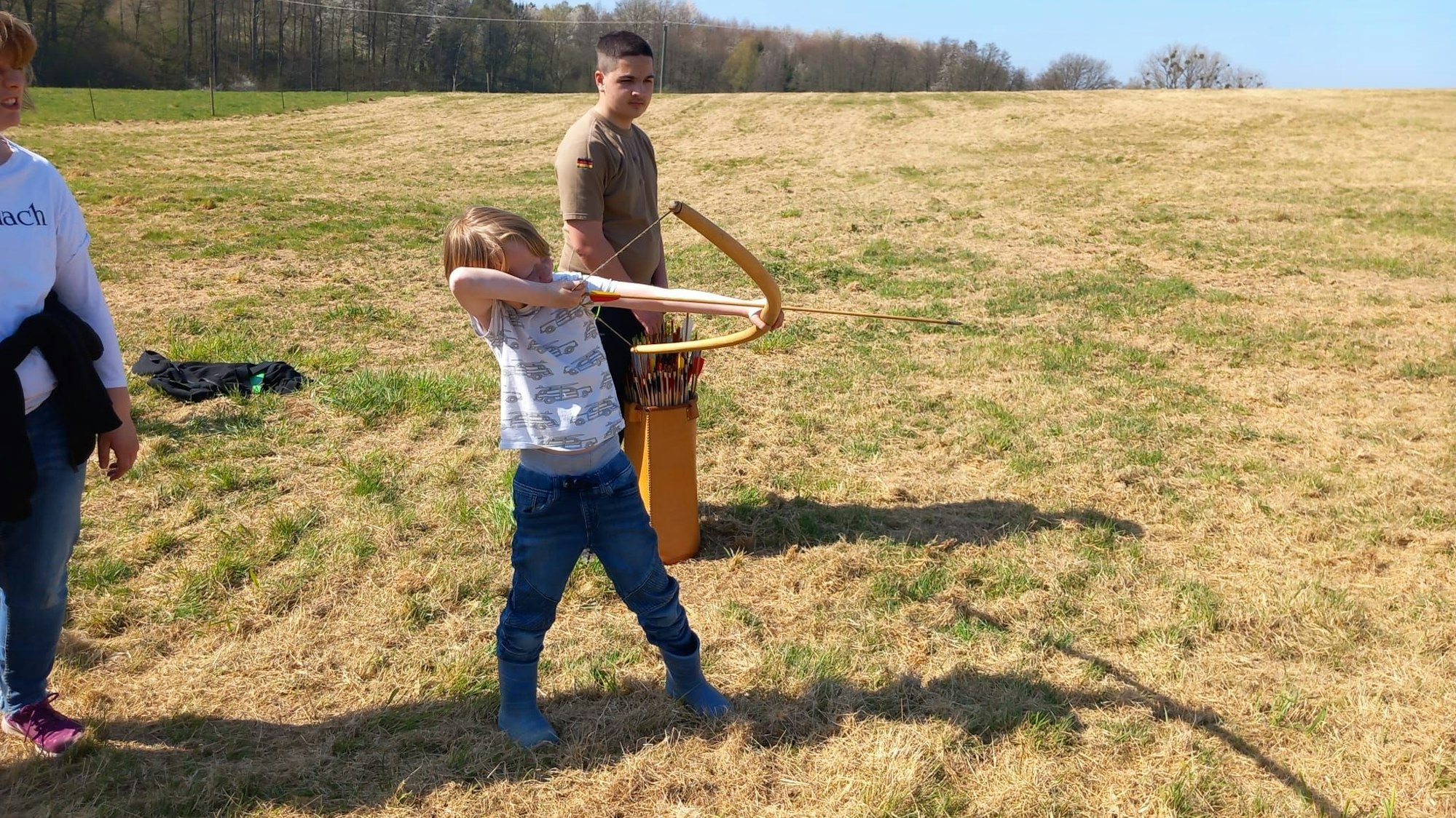 Ein Kind zielt mit einem Holzbogen auf einer Wiese.
