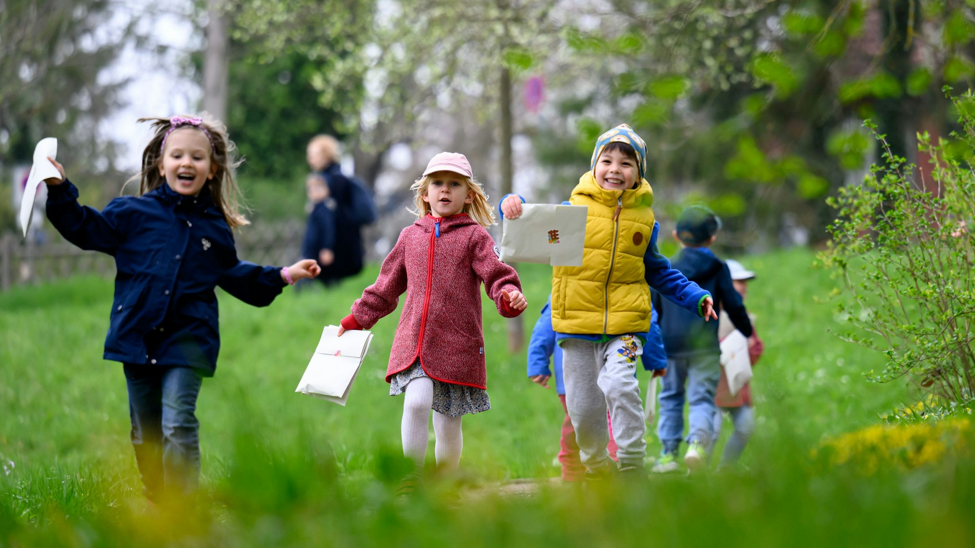 Kinder laufen mit Umschlägen in der Hand über eine Wiese, auf der Suche nach Ostereiern.