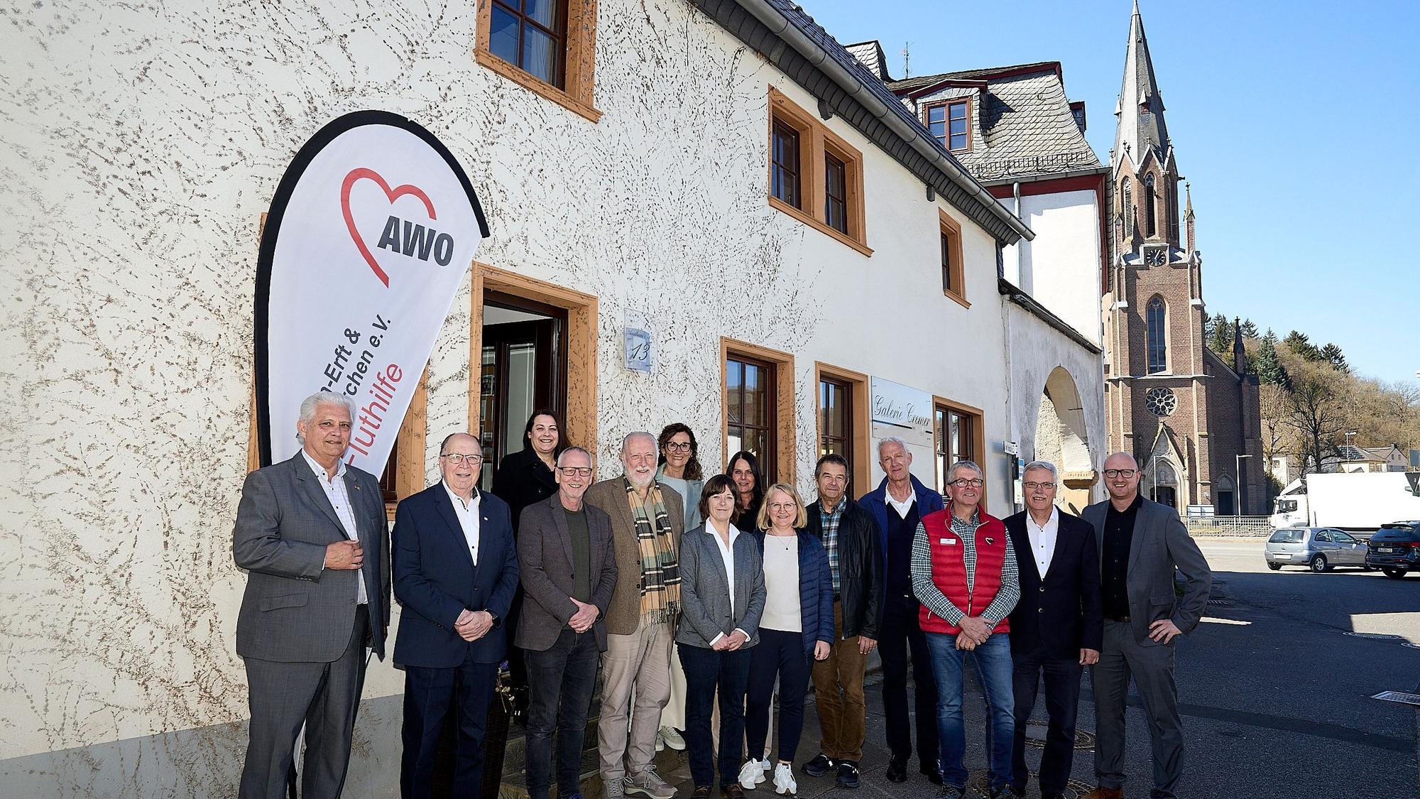Mehrere Menschen stehen vor einem Haus. Im Hintergrund sind Fahrzeuge und eine Kirche.